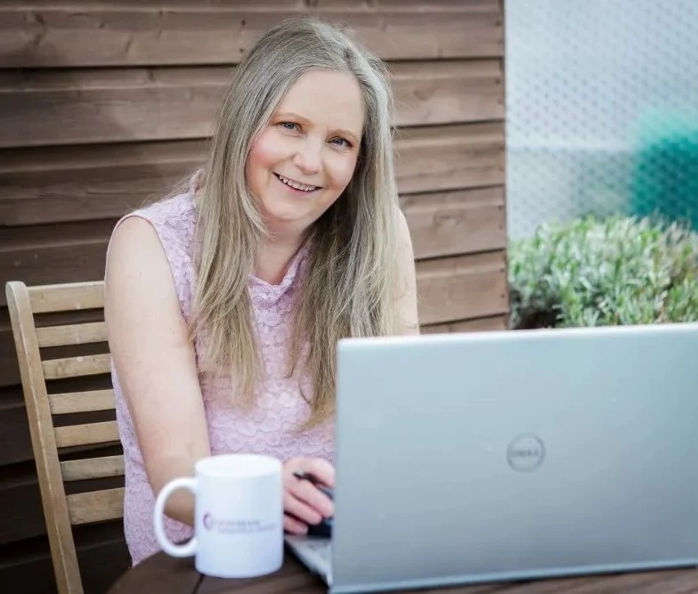 Karen, wearing a pink sleeveless sitting at wooden table in garden with silver laptop open and a white mug to the side of the laptop.